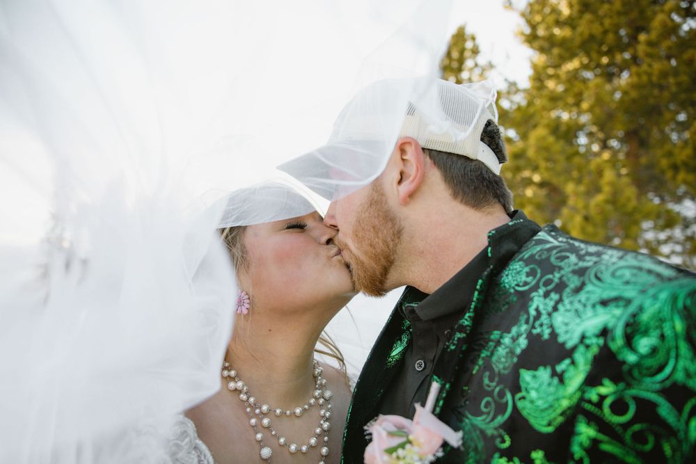 Bride and groom share a romantic kiss under a flowing white veil outdoors on their wedding day.