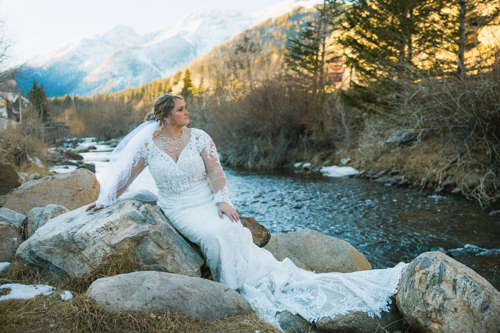 Bride in lace gown sits on rocks by mountain river, veil flowing, with snow-capped peaks in background.