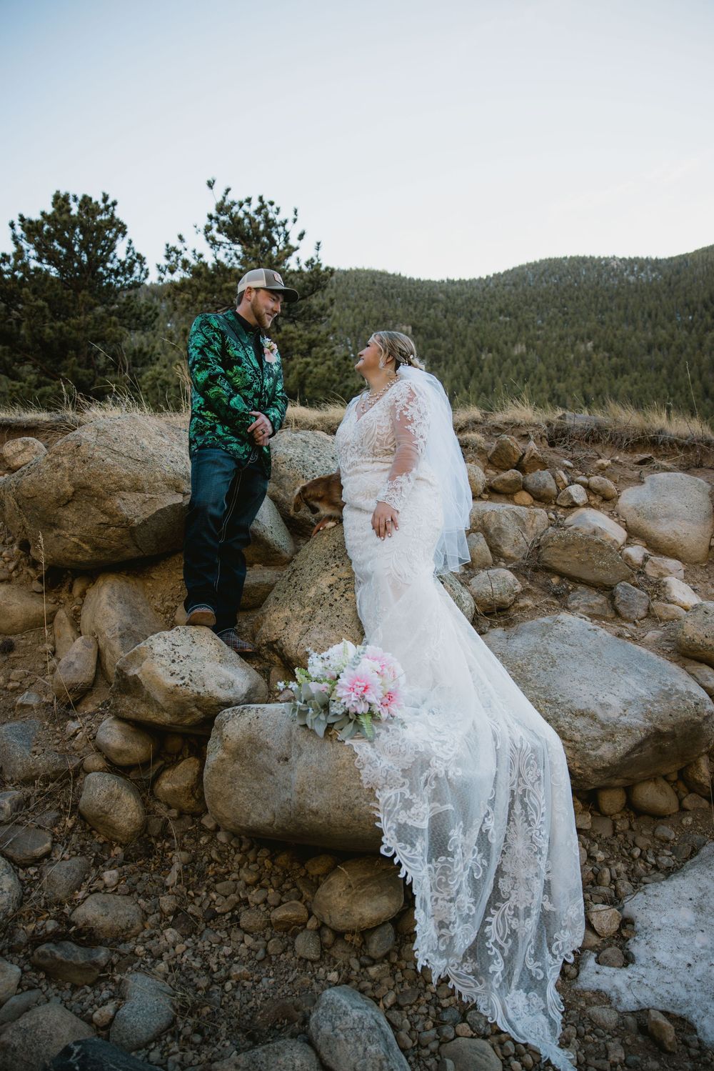 Bride in lace gown and groom in green jacket share a moment on rocky mountain terrain during outdoor elopement.