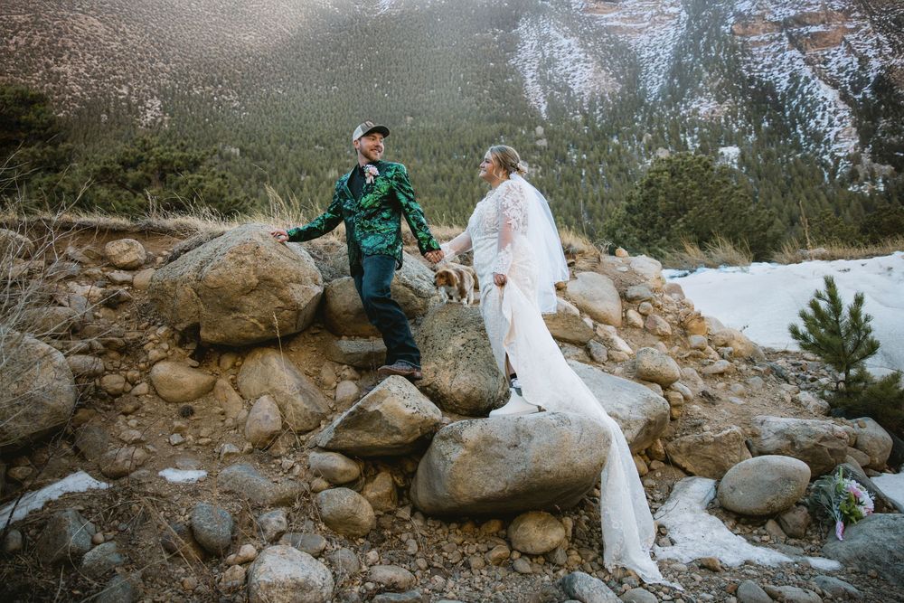Bride in white lace gown and groom in green jacket hold hands on rocky mountain terrain with snowy landscape backdrop.