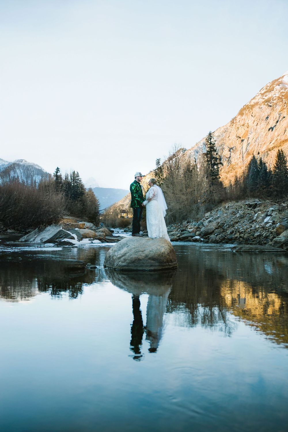 Bride and groom standing on a rock with their reflection in a calm mountain river.