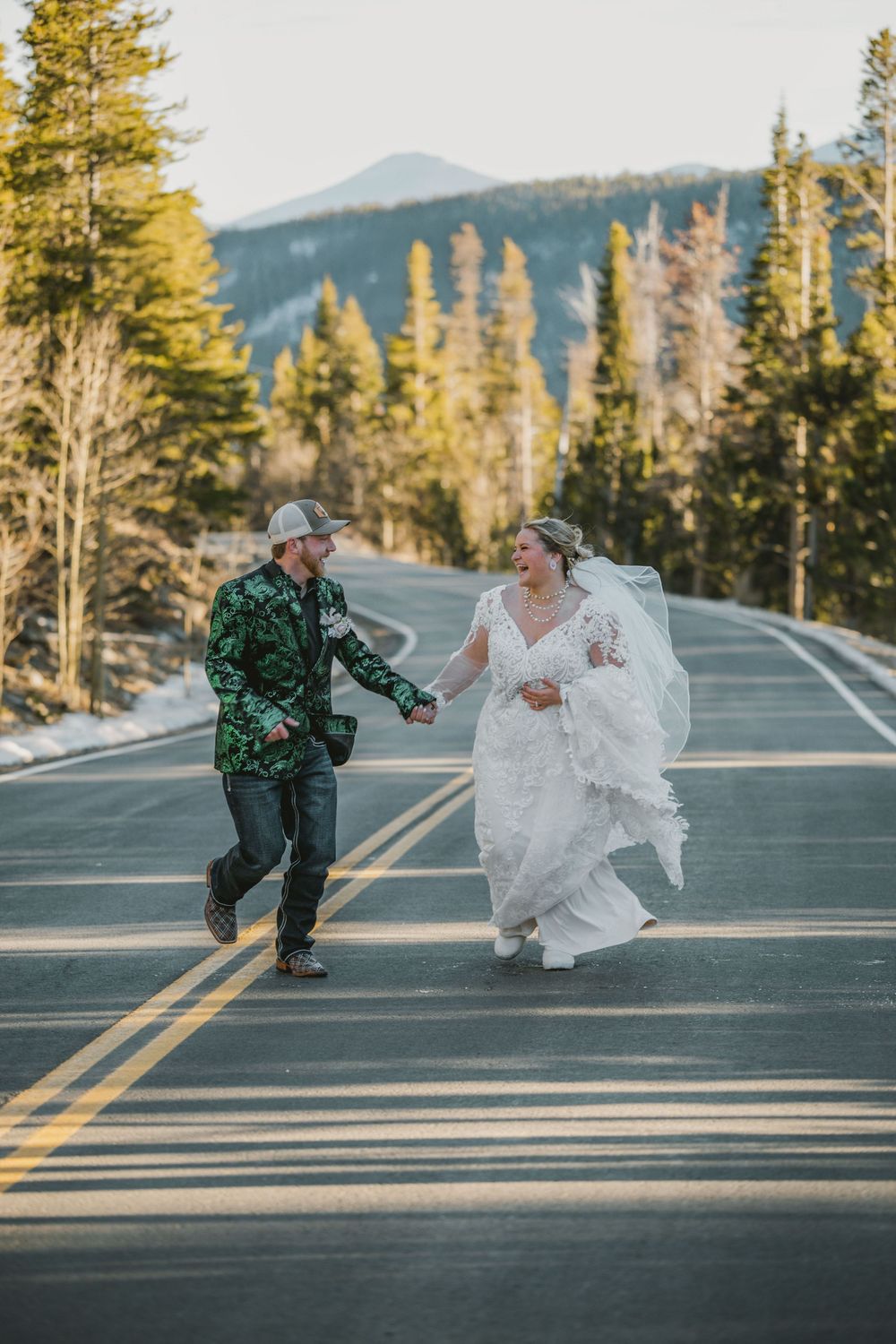 Bride and groom laughing while running on a mountain road surrounded by pine trees.