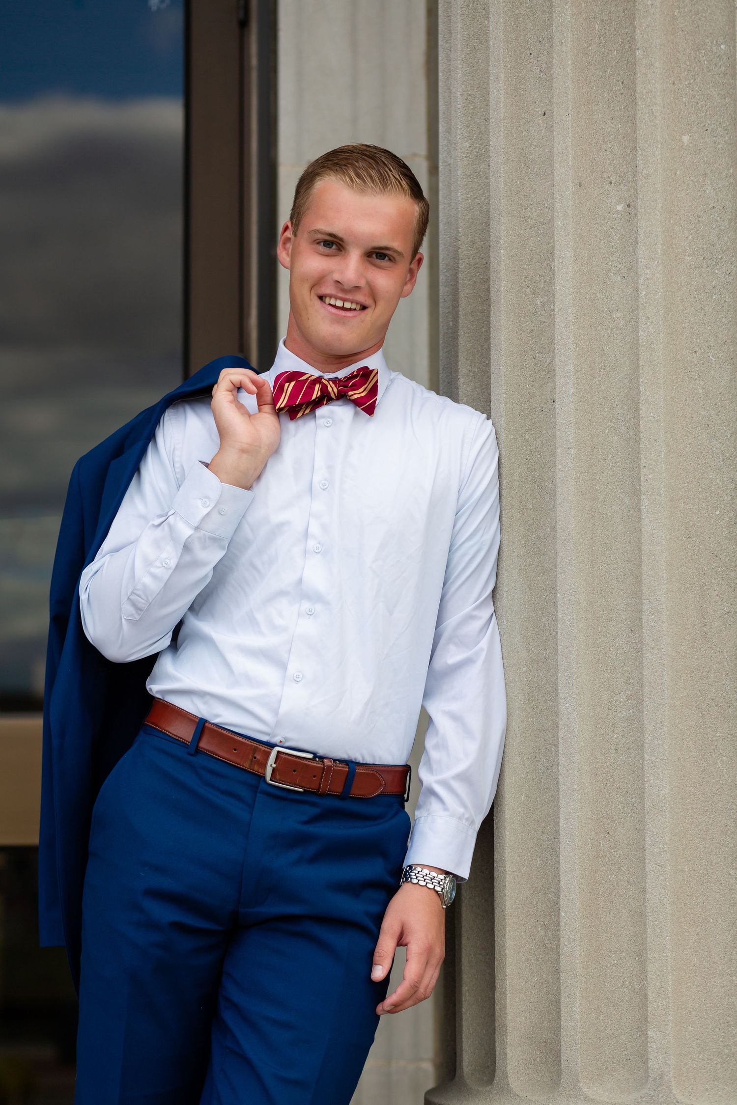 Young man in white shirt, red bow tie, and blue pants smiles, holding jacket over shoulder near stone column.