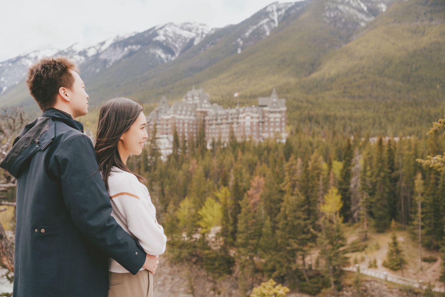 Couple gazing at Fairmont Banff Springs Hotel surrounded by pine trees and mountains in Banff, Canada.