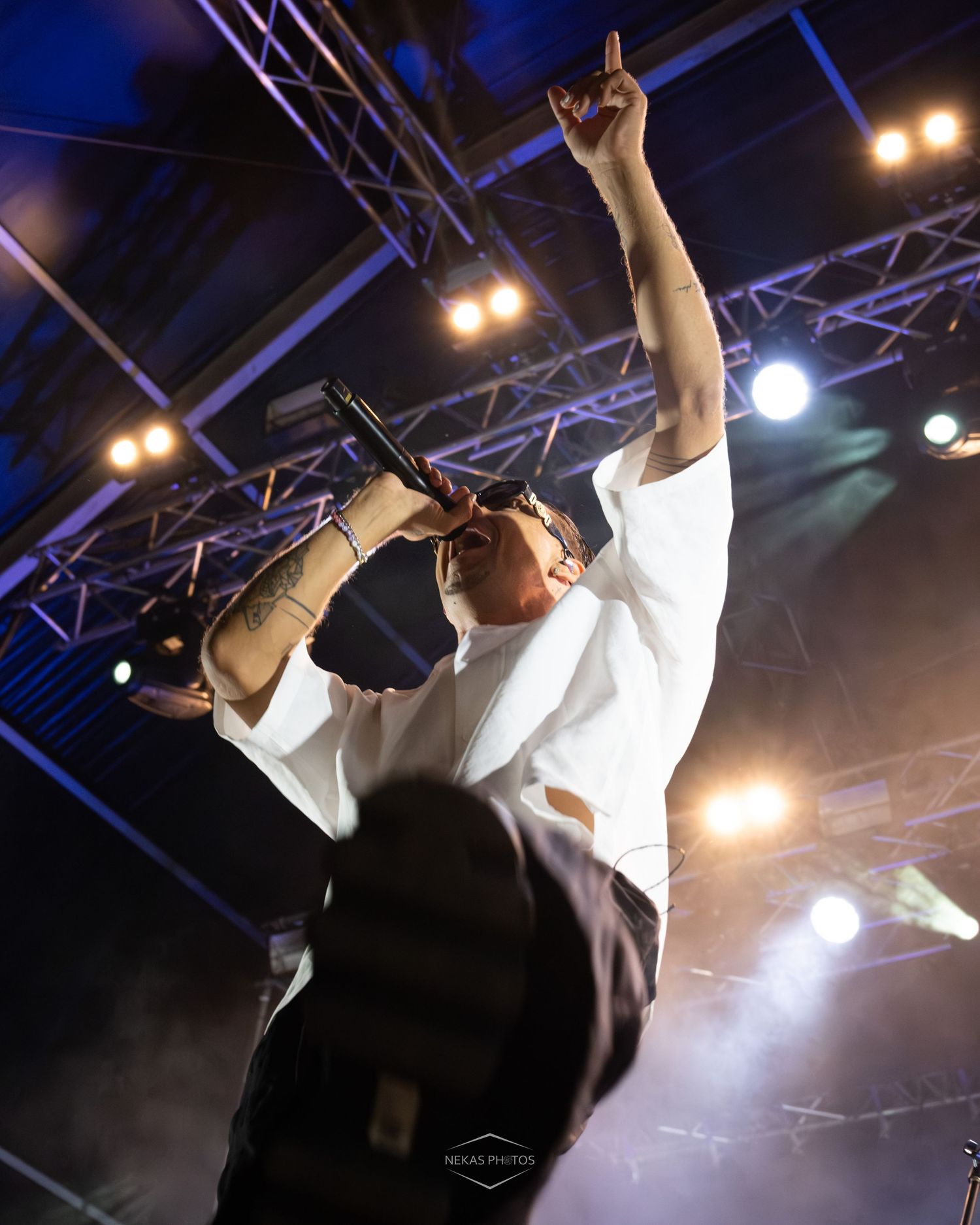 Performer in white shirt singing into microphone with one hand raised at an outdoor concert stage with colorful lights.