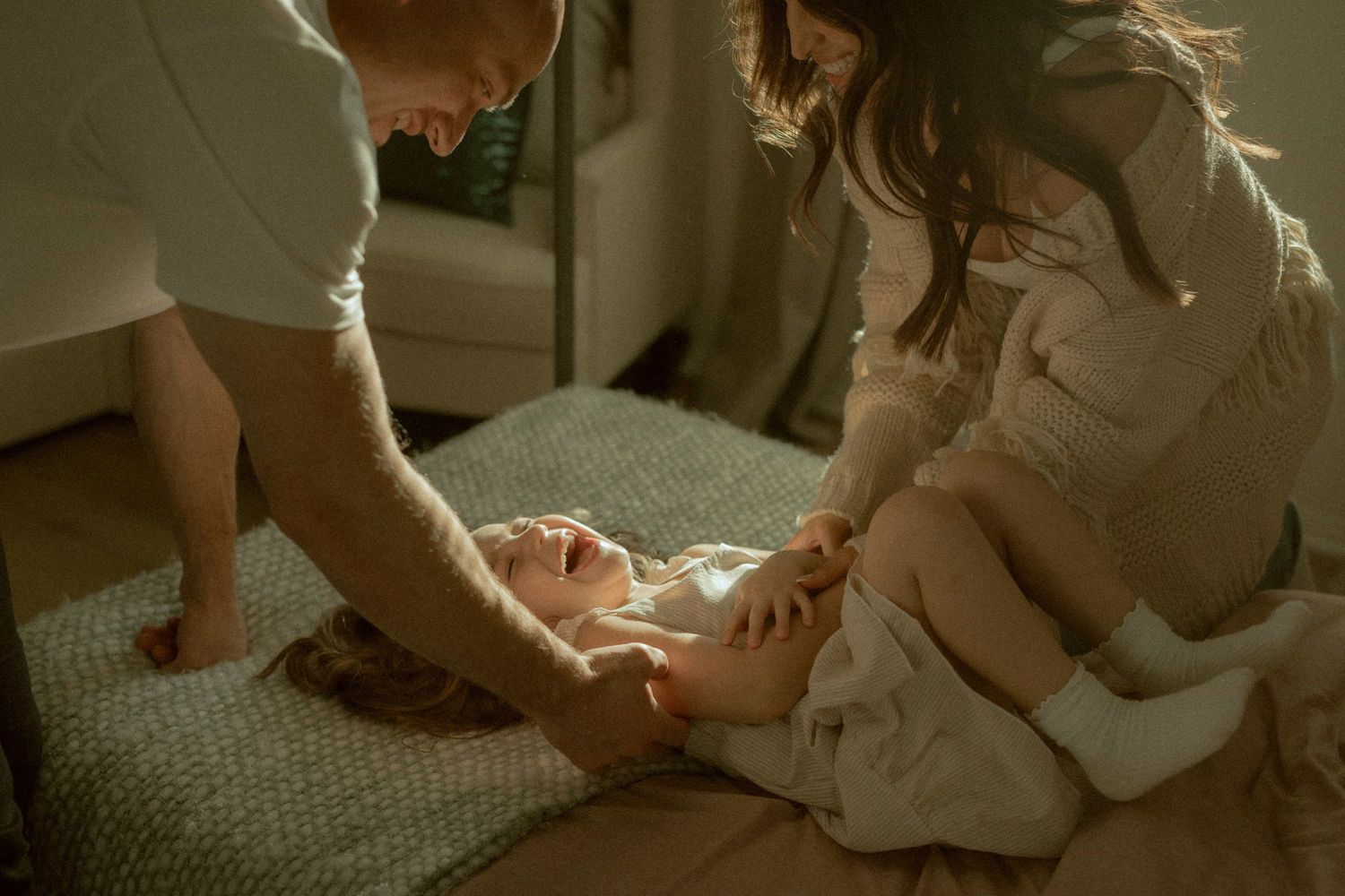 Parents playing with laughing baby on floor in warm natural indoor light.