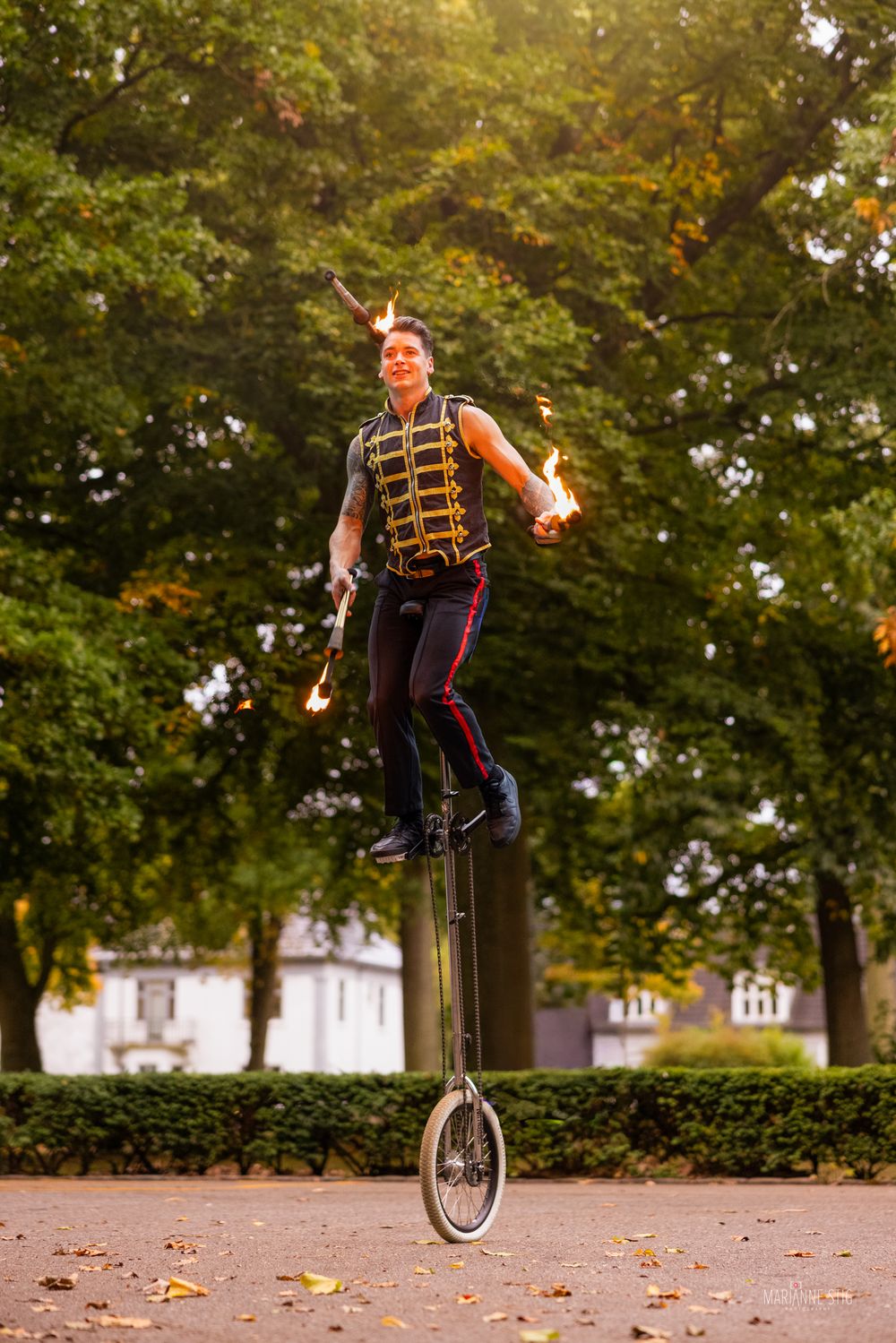 A performer juggles fire while balancing on a tall unicycle outdoors, wearing a decorative vest in a tree-lined park.