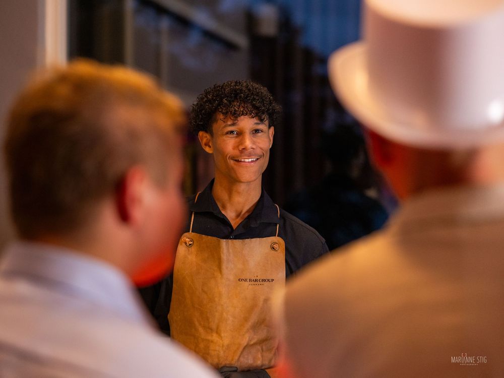 Young smiling waiter in brown apron greeting guests at an indoor event.