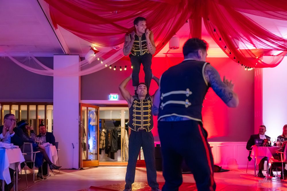 Circus performers in gold uniforms doing an acrobatic stunt at an indoor event with red draping decor.
