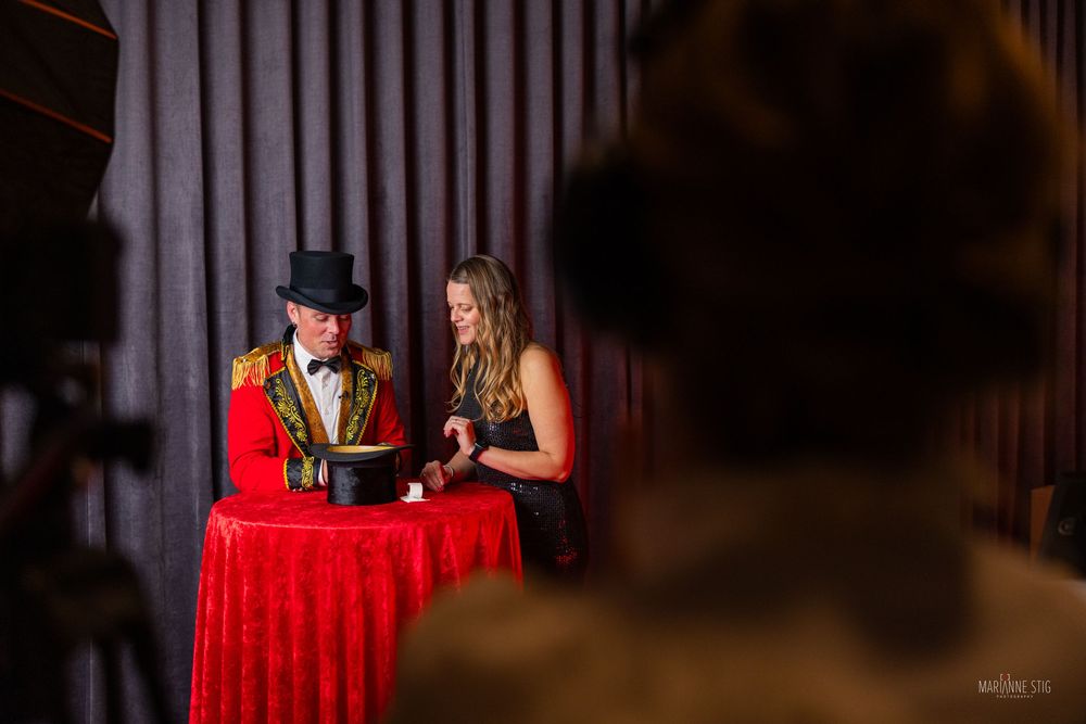 A magician in a top hat and woman in black dress examine a box on a red table at a circus-themed event.