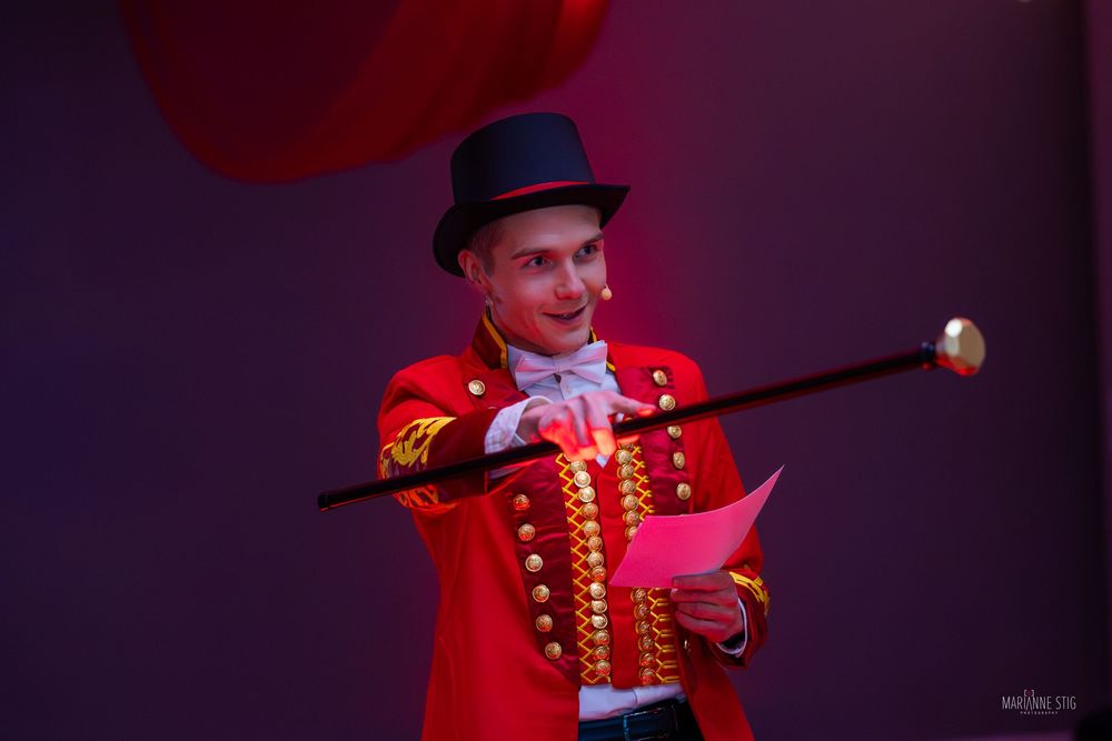 A smiling ringmaster in a red coat and black top hat holds a cane and pink card at a circus performance.