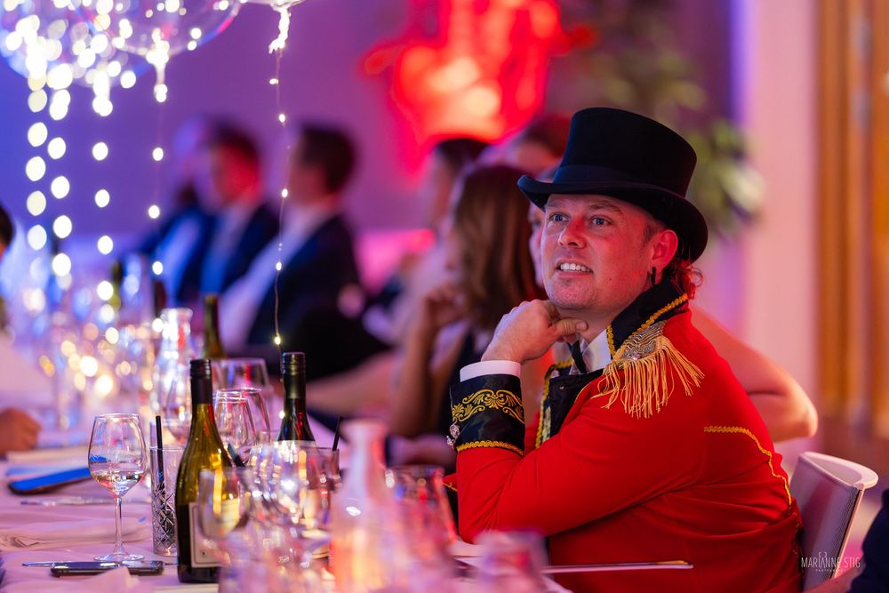 Man in red ringmaster costume and top hat seated at elegant gala dinner table with festive lighting.