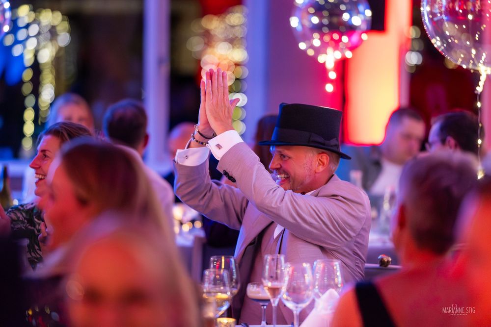 Man in black hat raises hands at elegant candlelit event with colorful lights in background.