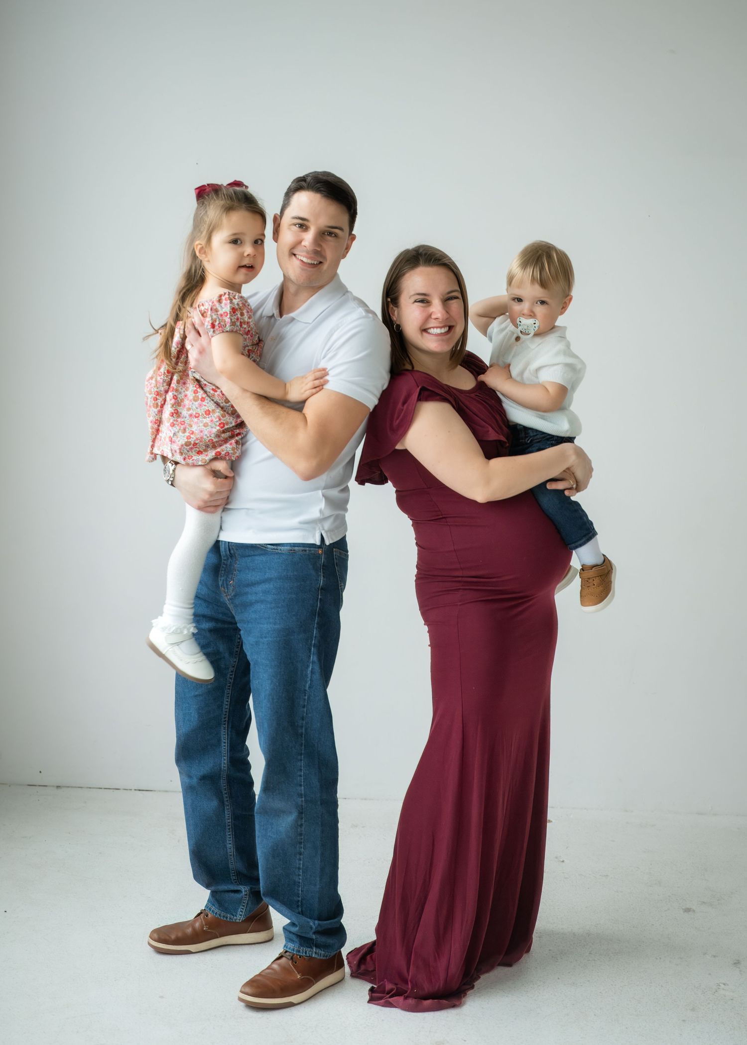Smiling pregnant woman in burgundy gown poses with husband and two young children in a bright studio maternity photo.