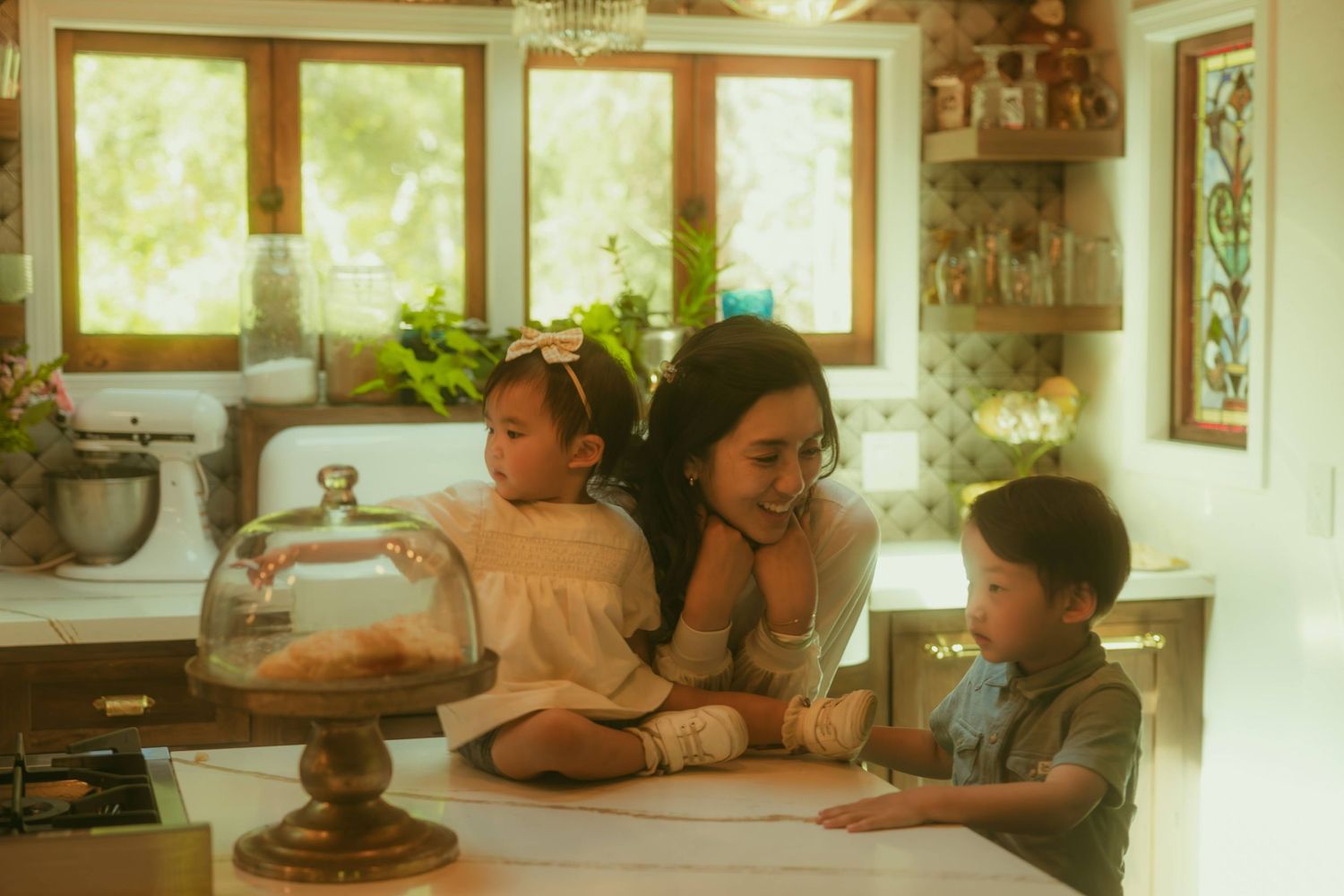 A mother smiles warmly with her two young children at a cozy kitchen counter in a bright, charming home.