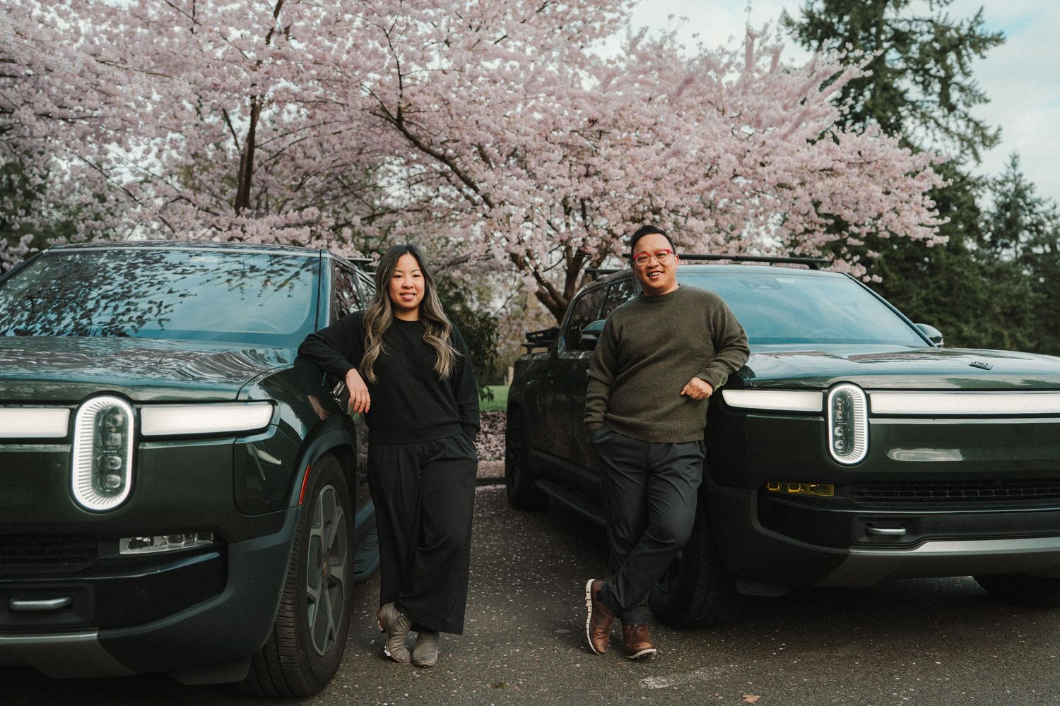 Two people pose between dark green Rivian electric trucks under blooming cherry blossom trees in spring.