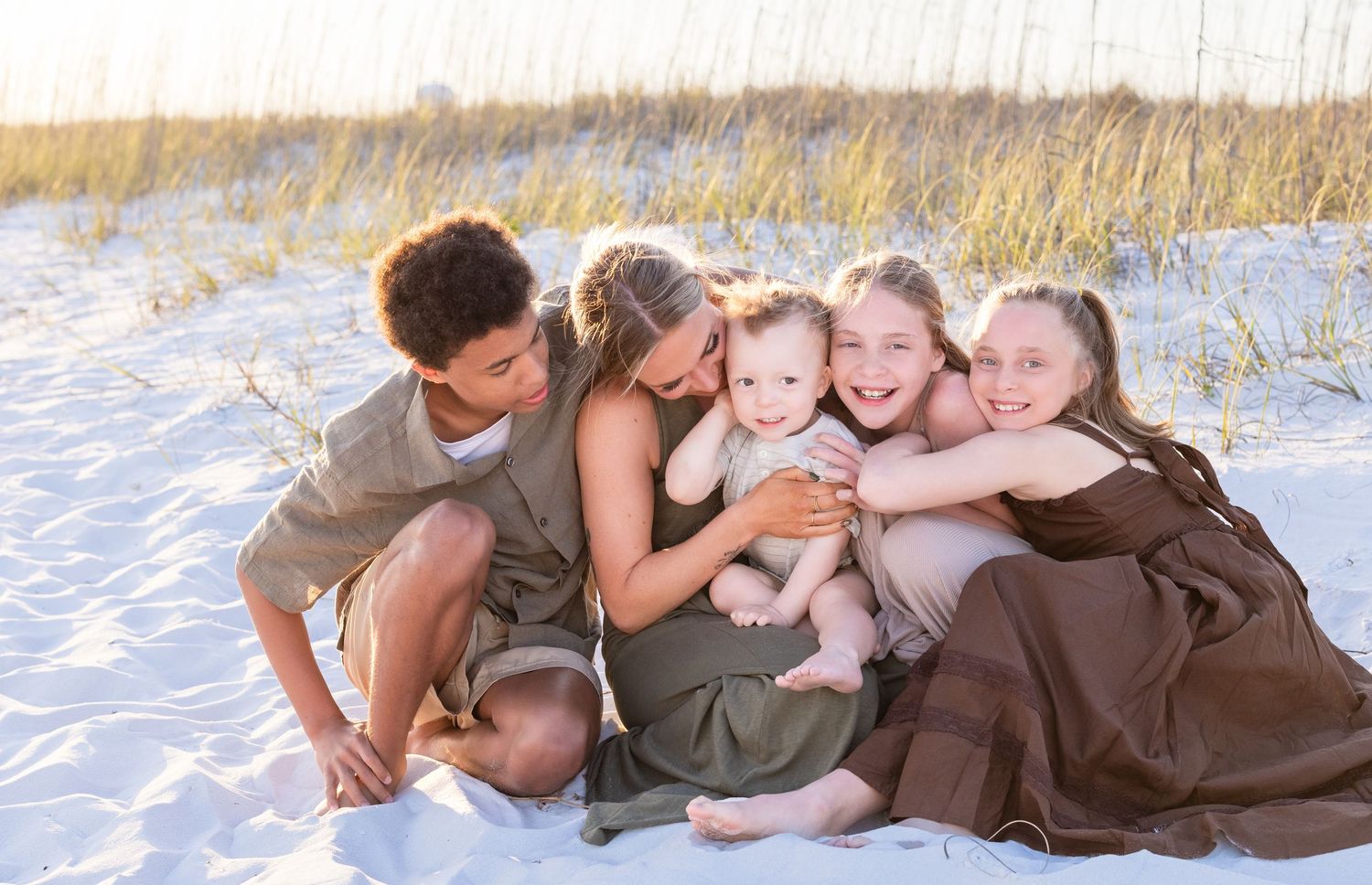 Family of five smiling together on white sand beach, children hugging mom in warm golden light.