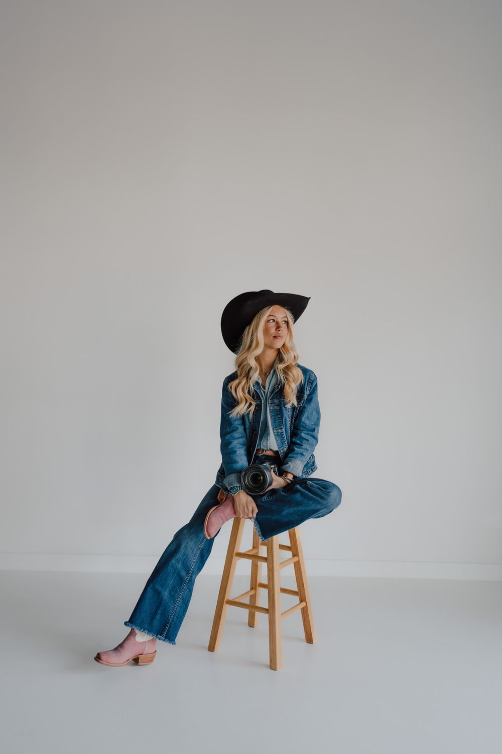 Smiling blonde woman in denim jacket and black cowboy hat sitting on stool holding a camera in a bright studio.
