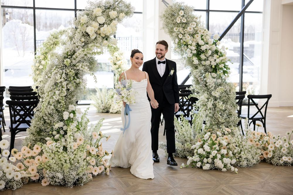 Bride and groom smiling under elegant white floral arch with roses and greenery at indoor wedding.