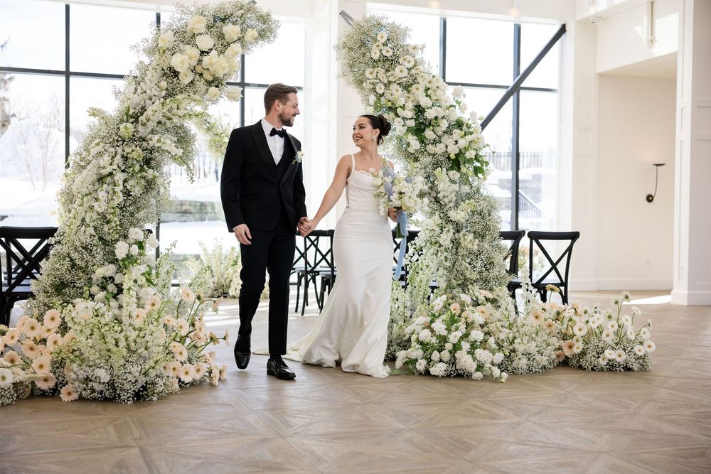 Newlyweds holding hands beside stunning double white floral arch at bright indoor wedding venue.