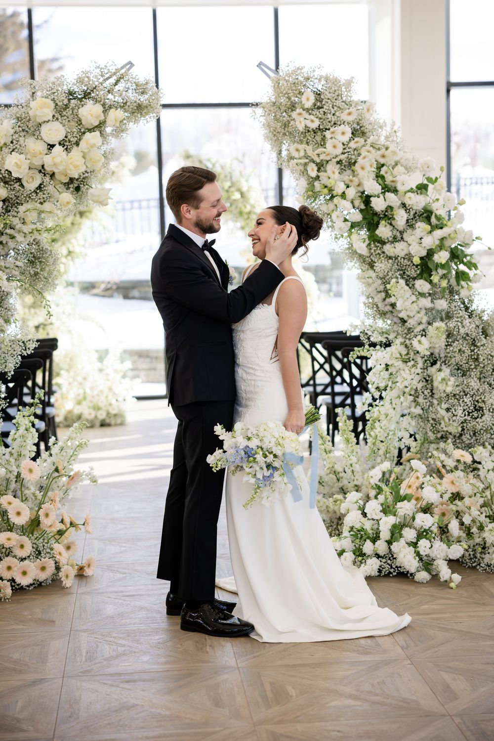 Groom tenderly touching bride's face under lush white rose and baby's breath wedding floral arch.