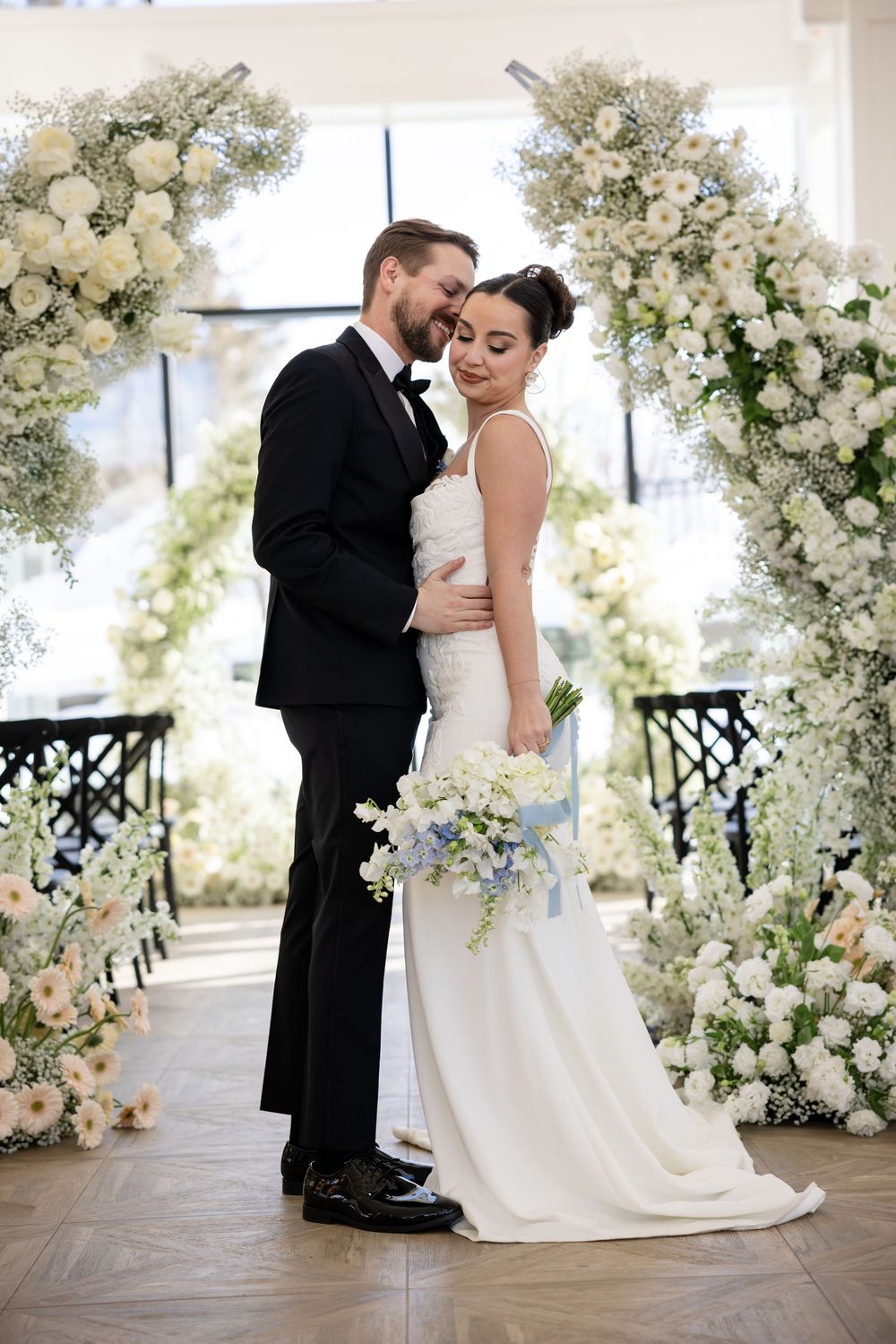 Bride and groom embracing with blue and white bouquet before grand white floral arch at wedding.
