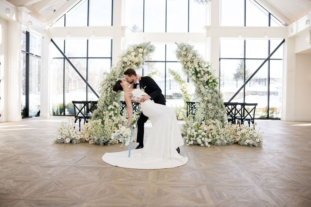 Groom dramatically dipping bride in kiss under white floral arch in bright elegant wedding venue.