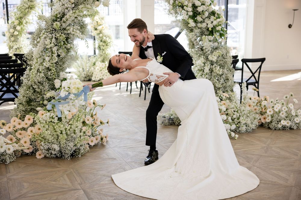 Groom dipping bride in romantic pose before stunning white and pink floral arch at wedding ceremony.