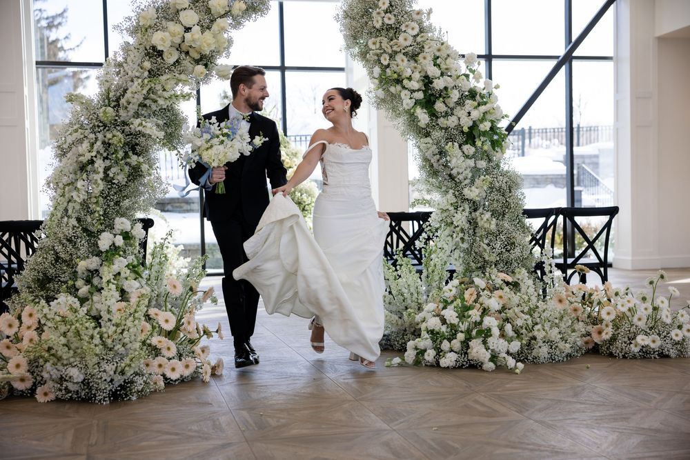 Joyful bride and groom walking under white floral arch in bright elegant indoor wedding venue.