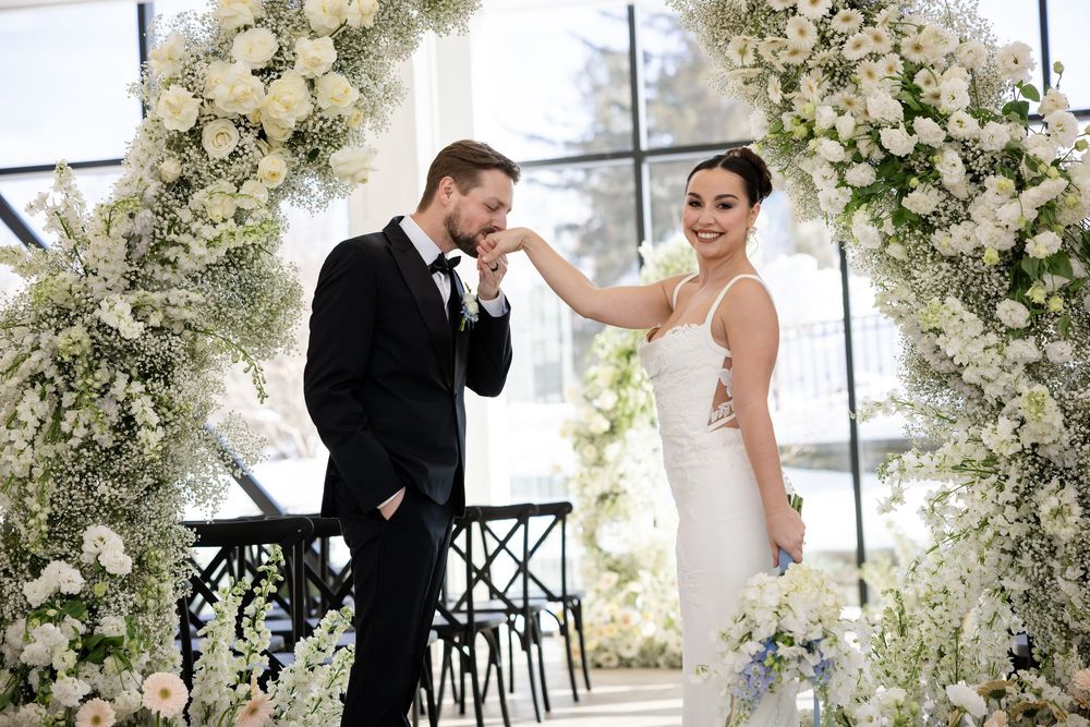 Groom kissing bride's hand under white floral arch at elegant indoor wedding ceremony.