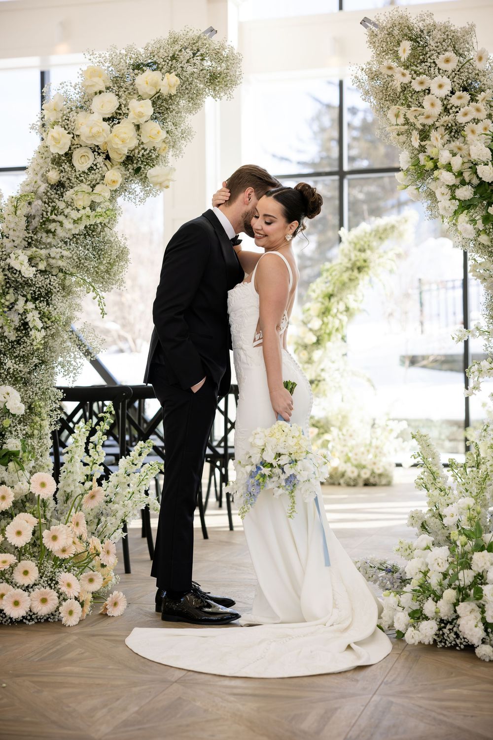 Bride and groom sharing romantic moment under lush white flower arch at indoor wedding.