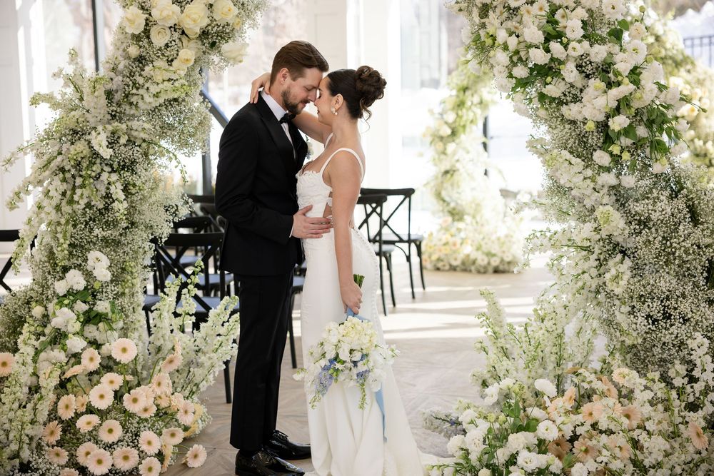 Bride and groom sharing intimate forehead touch under lush white floral arch at elegant wedding.