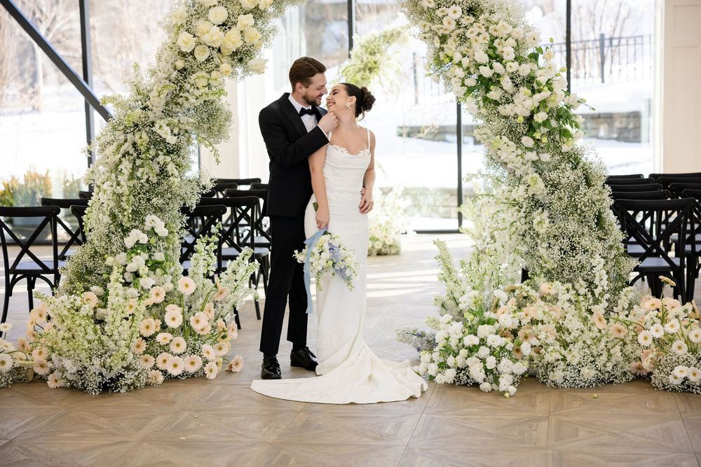Couple sharing a kiss beneath a stunning white floral arch at an indoor wedding venue.
