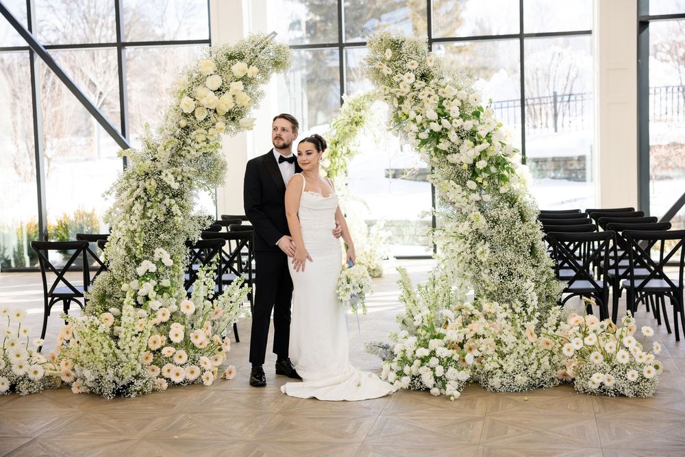 Bride and groom posing together under an elaborate white floral arch at indoor ceremony.