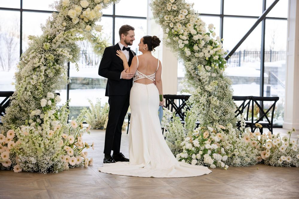 Bride in backless gown and groom standing before white floral arch at wedding ceremony.