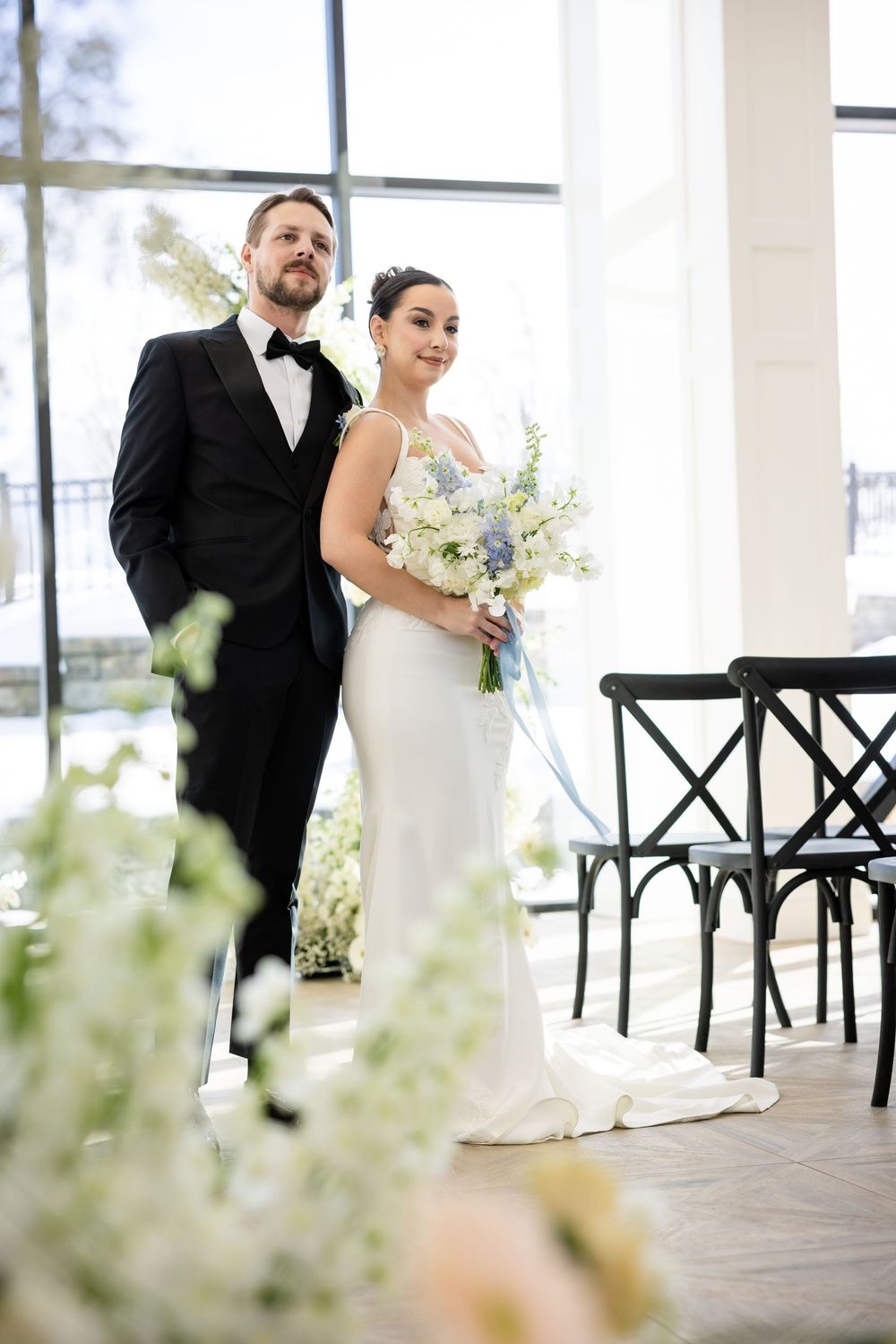 Bride holding blue and white bouquet standing with groom in bright windowed wedding venue.
