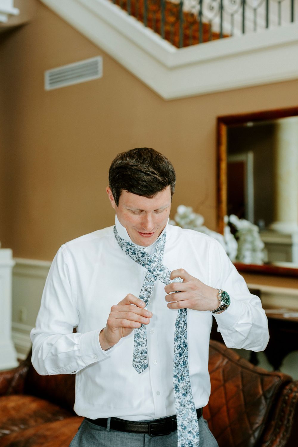 Man in white dress shirt tying a floral patterned necktie while getting ready indoors near a staircase.