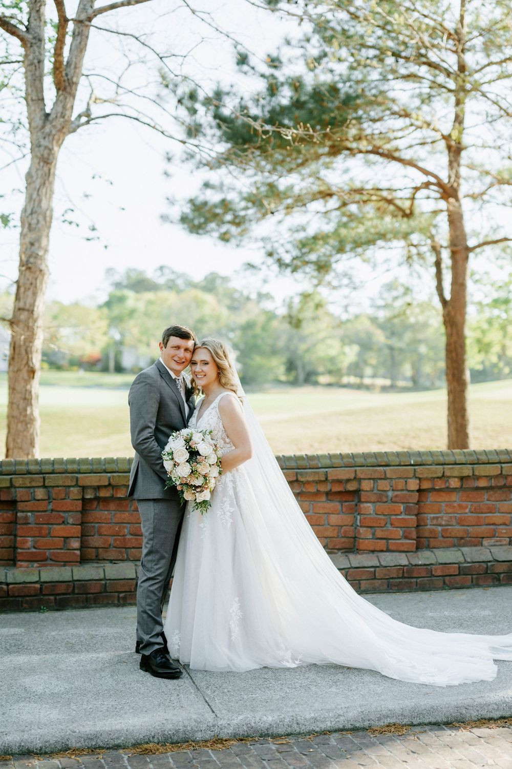 Bride and groom pose together outdoors near a brick wall, surrounded by tall trees and lush greenery on their wedding day.