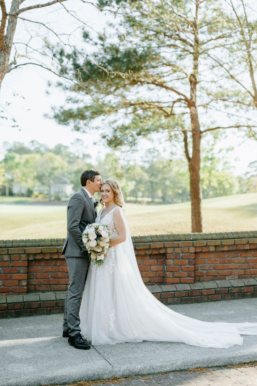 Bride in white gown holding bouquet stands with groom in gray suit near brick wall and pine trees at outdoor wedding.
