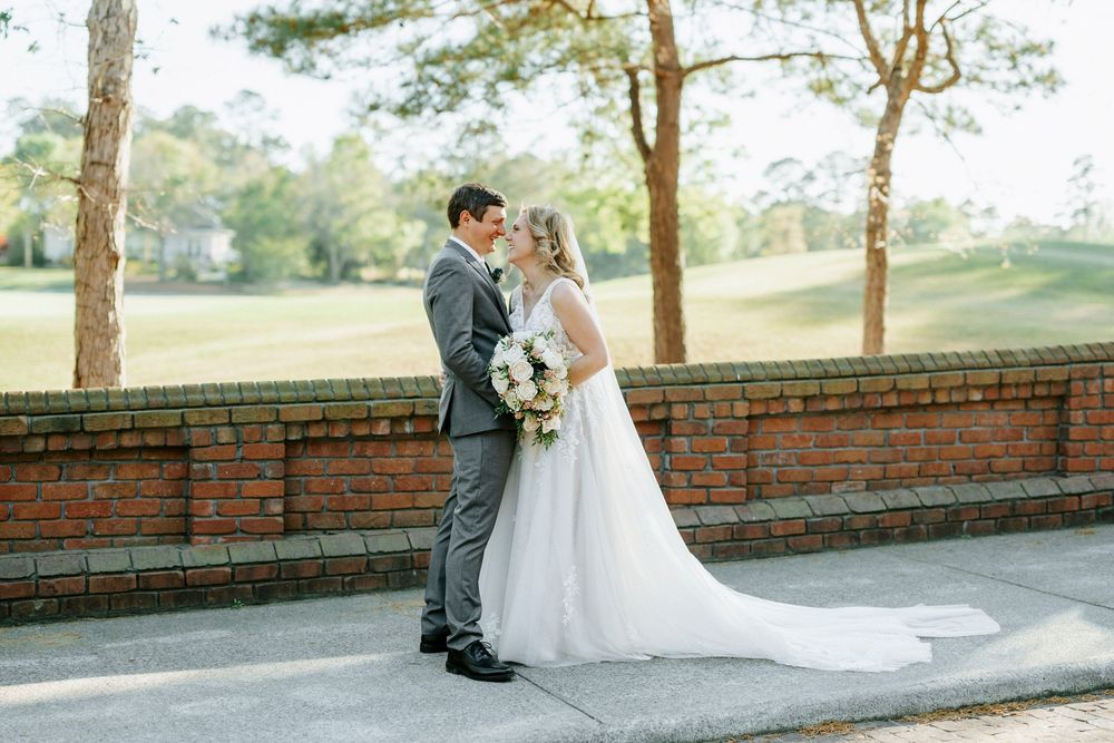 Bride and groom share a romantic moment outdoors near a brick wall, with lush greenery and trees in the background.