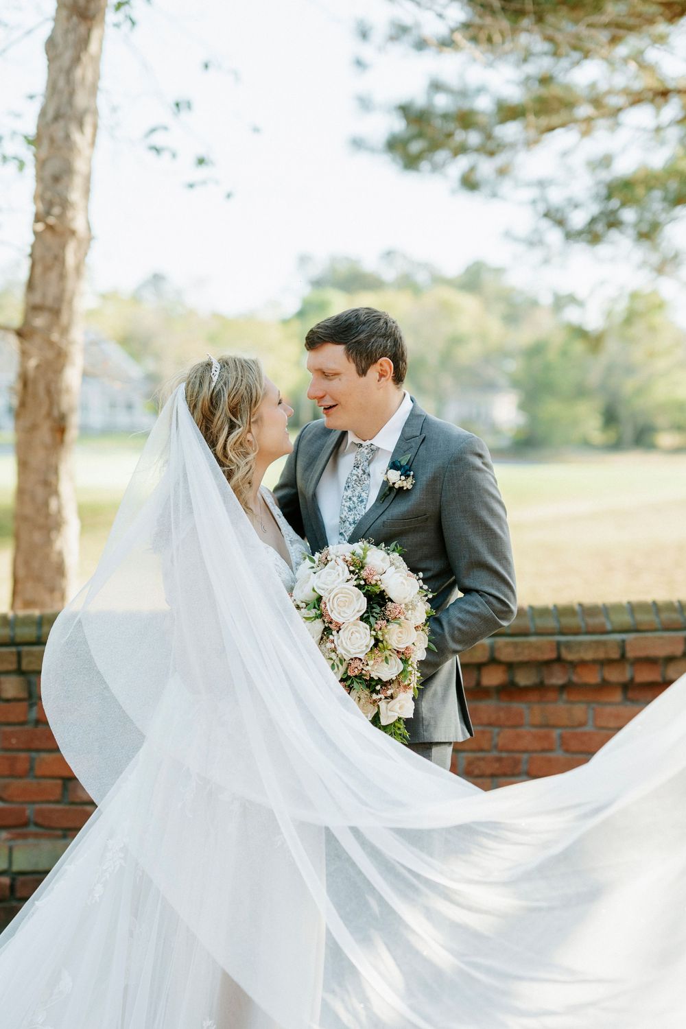 Bride and groom share a joyful moment outdoors, her veil flowing in the wind, holding a pink bouquet.