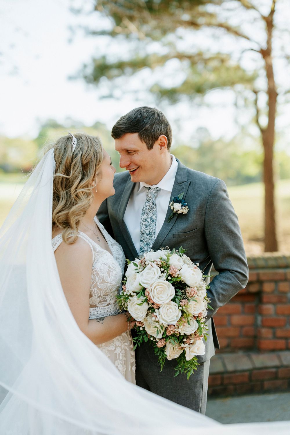 Bride and groom share a romantic moment outdoors, bride holding a bouquet of blush roses, groom in a gray suit.
