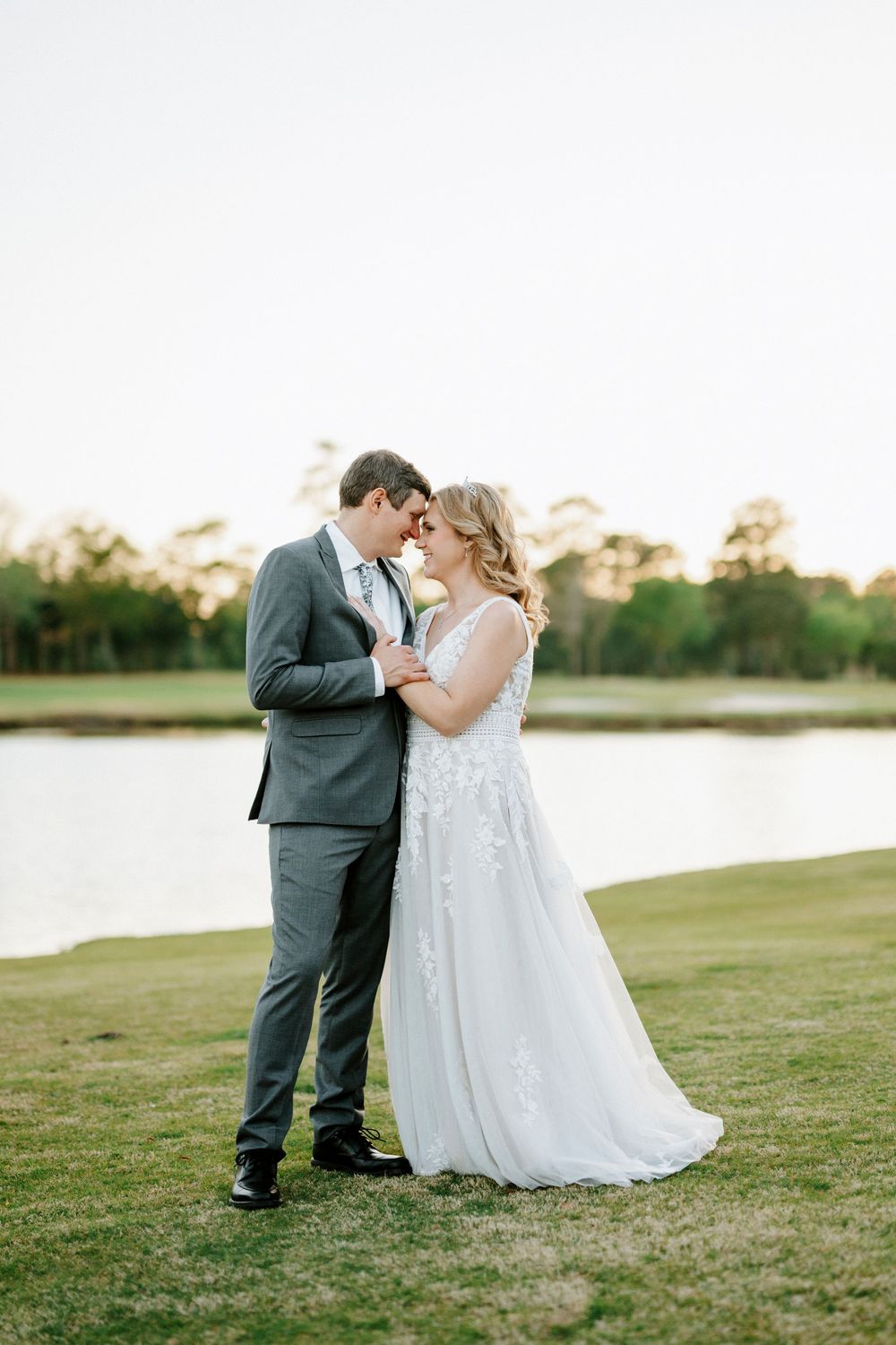 A bride and groom share a romantic forehead touch in a lush outdoor setting near a serene lake at golden hour.