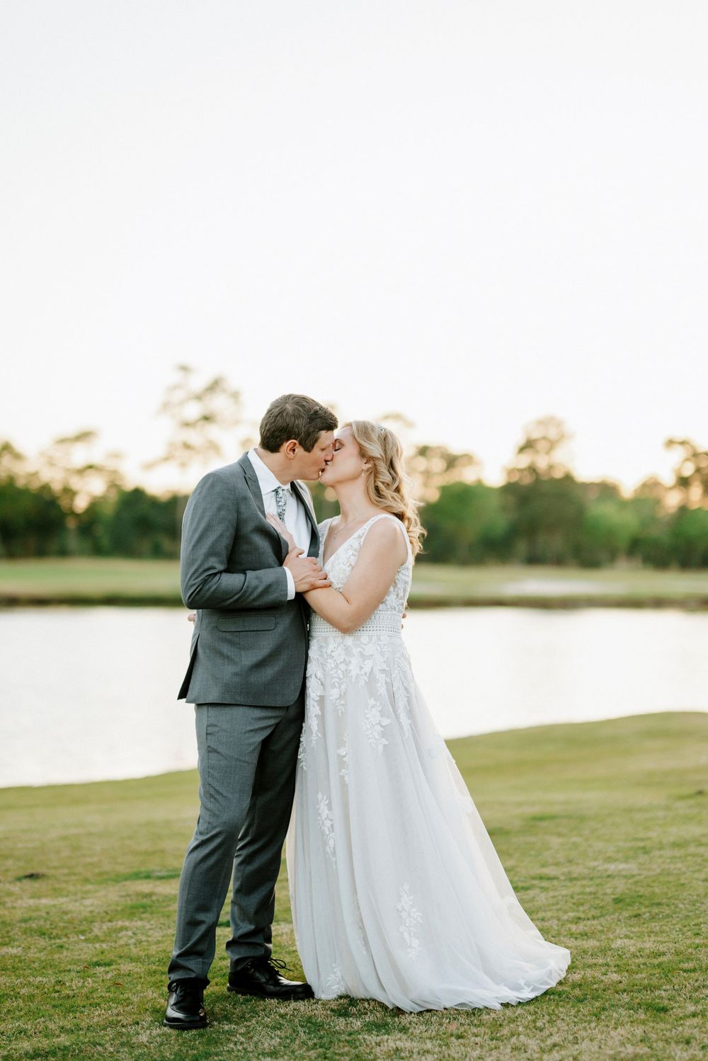 Bride and groom share a romantic kiss in front of a serene pond at golden hour during their outdoor wedding.