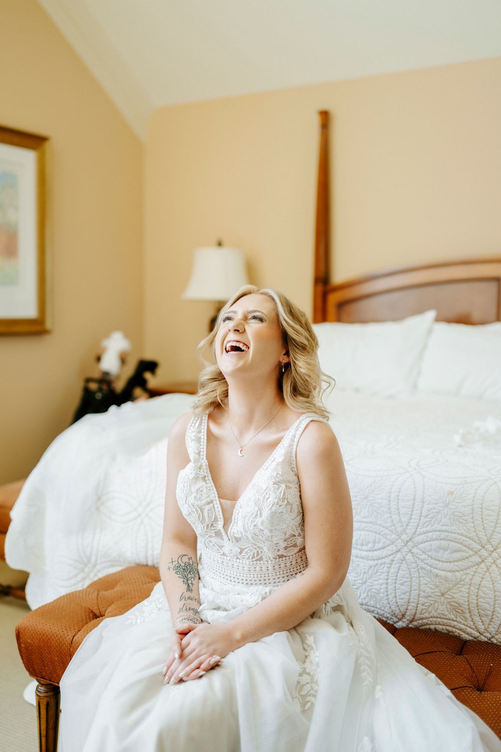 Laughing bride in white lace wedding gown sits on bed bench in warm hotel room, radiating joy on her wedding day.