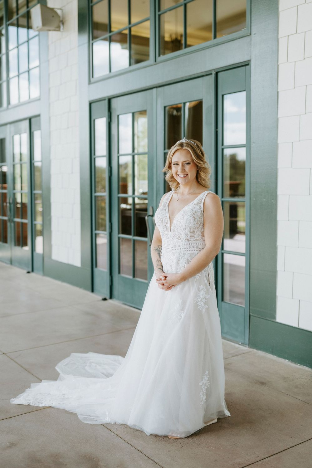 Smiling bride in a white lace A-line wedding gown with a train, posing in front of green-framed glass doors.