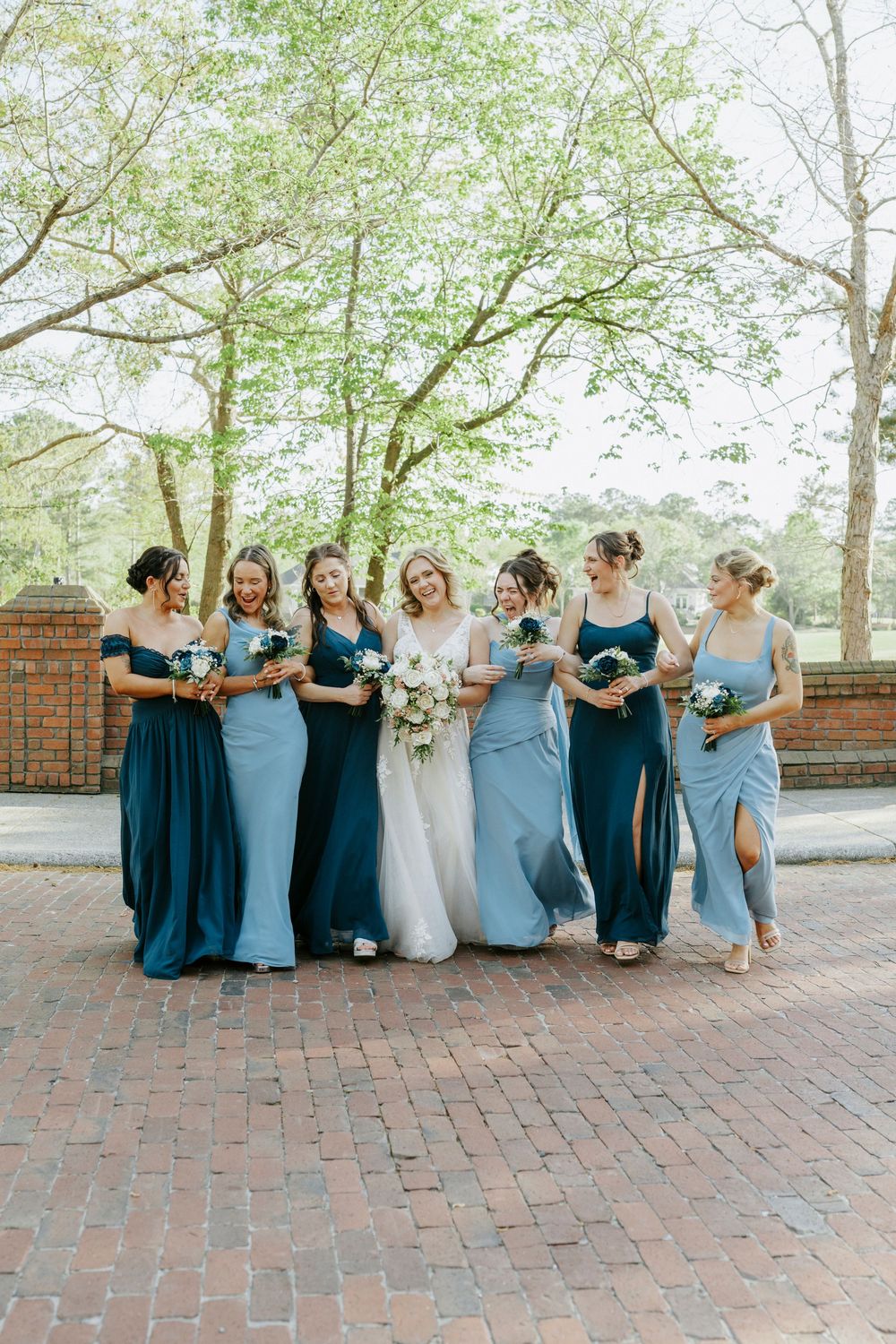 Bride walks with five bridesmaids in blue gowns holding bouquets on a brick path surrounded by trees.