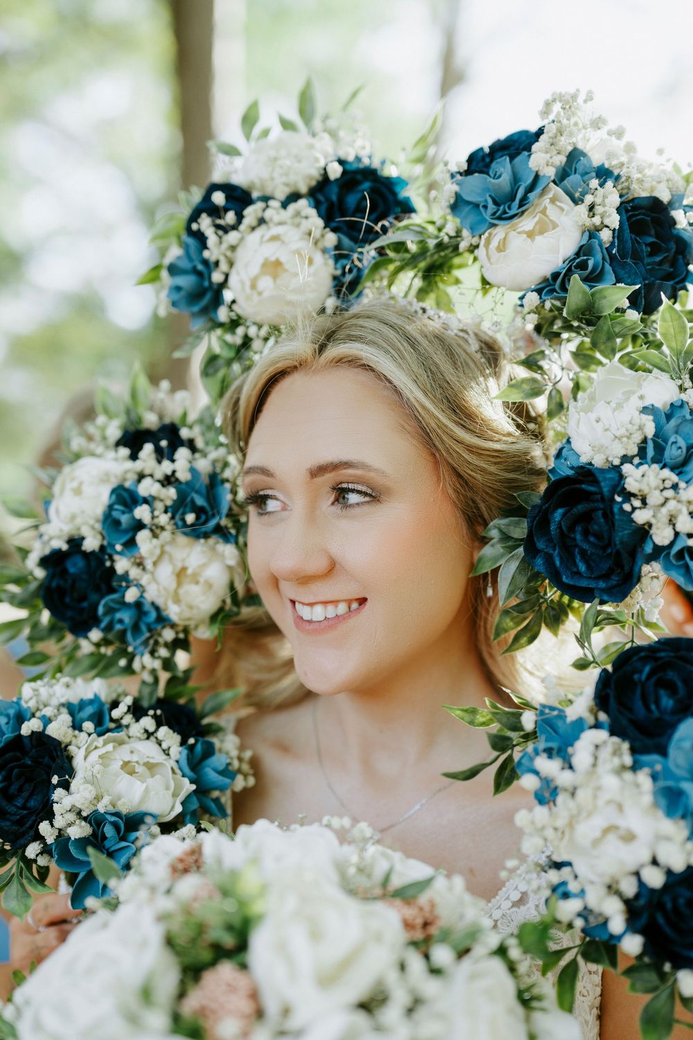 Smiling bride surrounded by blue and white floral bouquets with roses and greenery on her wedding day.