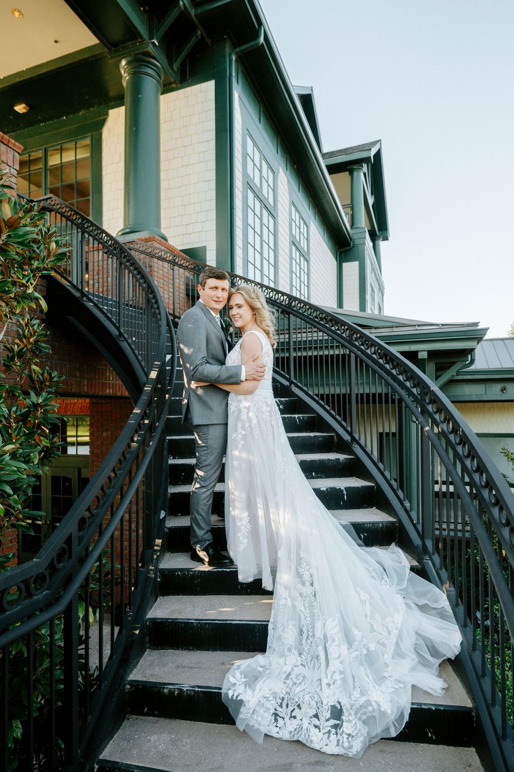 Bride and groom pose on elegant curved black iron staircase outside historic building in wedding attire with lace train.