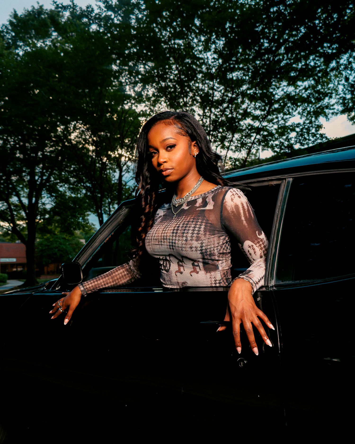 Young woman in sheer patterned top poses beside black car outdoors at dusk with green trees in background.